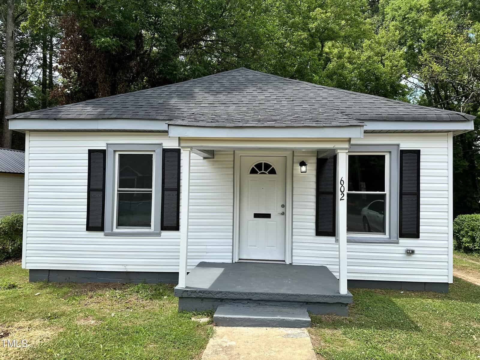 A small white house with dark shutters and a dark roof, featuring a central front door with an arch window. The house has a covered front porch with three steps leading up to it. The number "602" is displayed beside the door. Lush green trees stand behind the house.