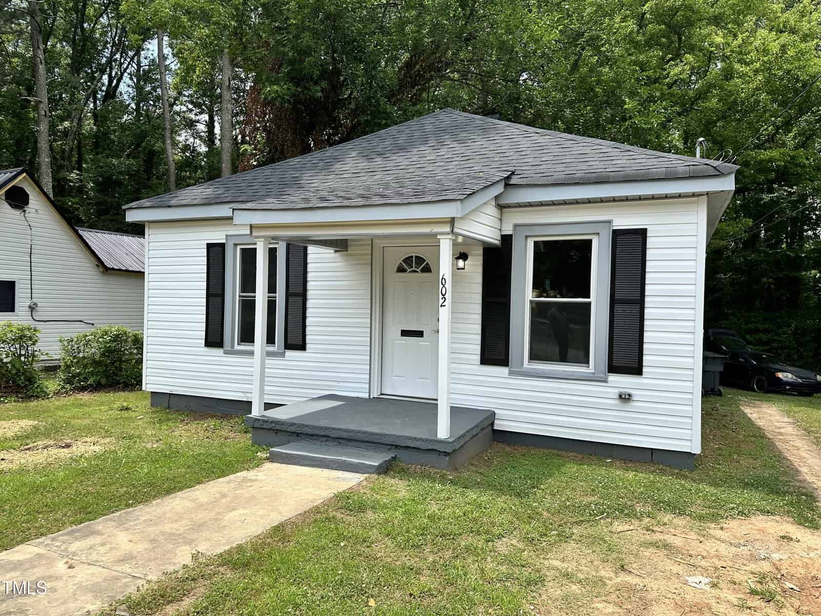 A small white house with a gray shingled roof and black shutters sits quaintly at 602 High Street in Henderson, NC. It features a small porch with two steps leading to a white door with an arched window at the top. A concrete walkway graces the front yard, and trees are visible in the background. House number "502" is visible.