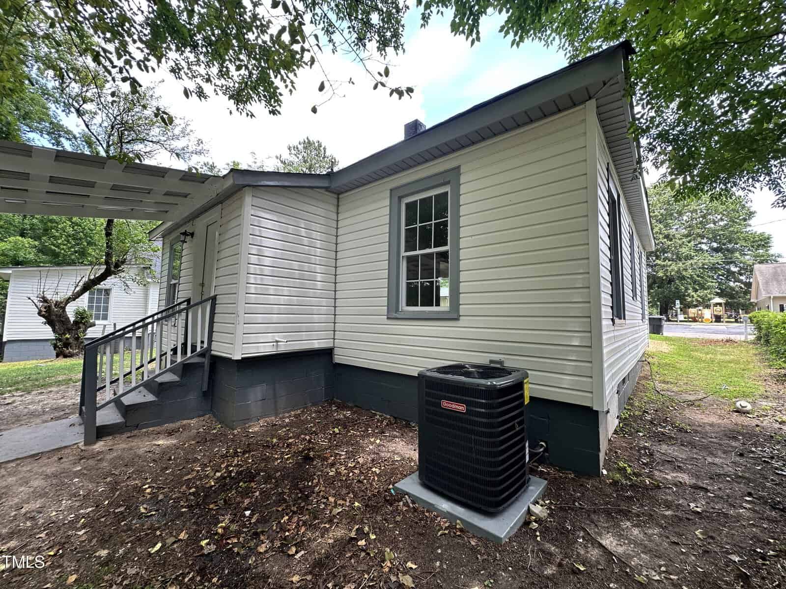 A small white house with gray trim sits on a grassy yard with some dirt patches. It has a covered side porch with steps leading up. A black air conditioning unit is situated on one side of the house. Trees and another house are visible in the background.