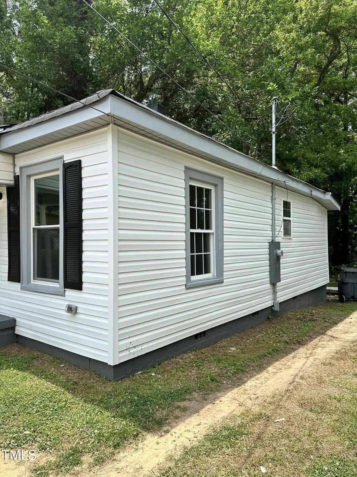 A small white house with vinyl siding, black shutters, and a gray roof. It has a grassy yard and is surrounded by trees. An electrical meter and wires are attached to the side of the house. The house windows have white trims.