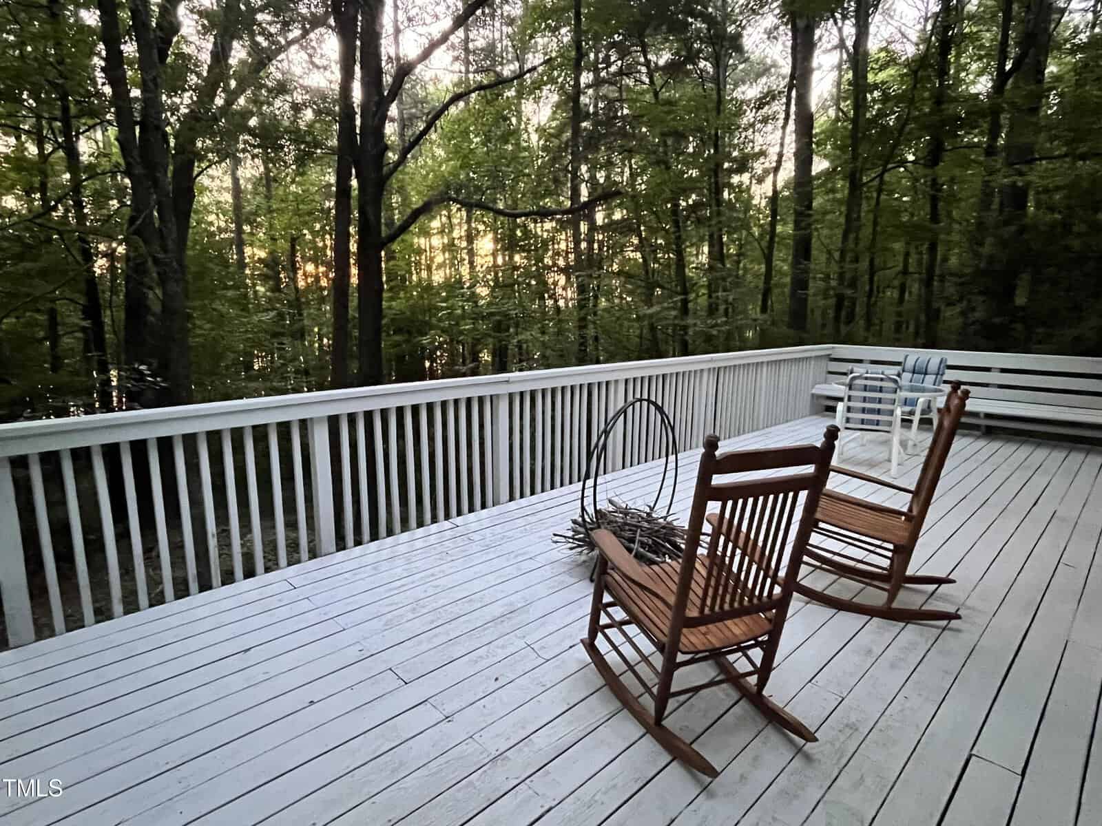 A wooden deck surrounded by trees features two wooden rocking chairs and a circular fire pit on the white-painted floor. In the background, a bench and a striped towel are visible. Sunlight filters through the dense foliage, creating a peaceful atmosphere.