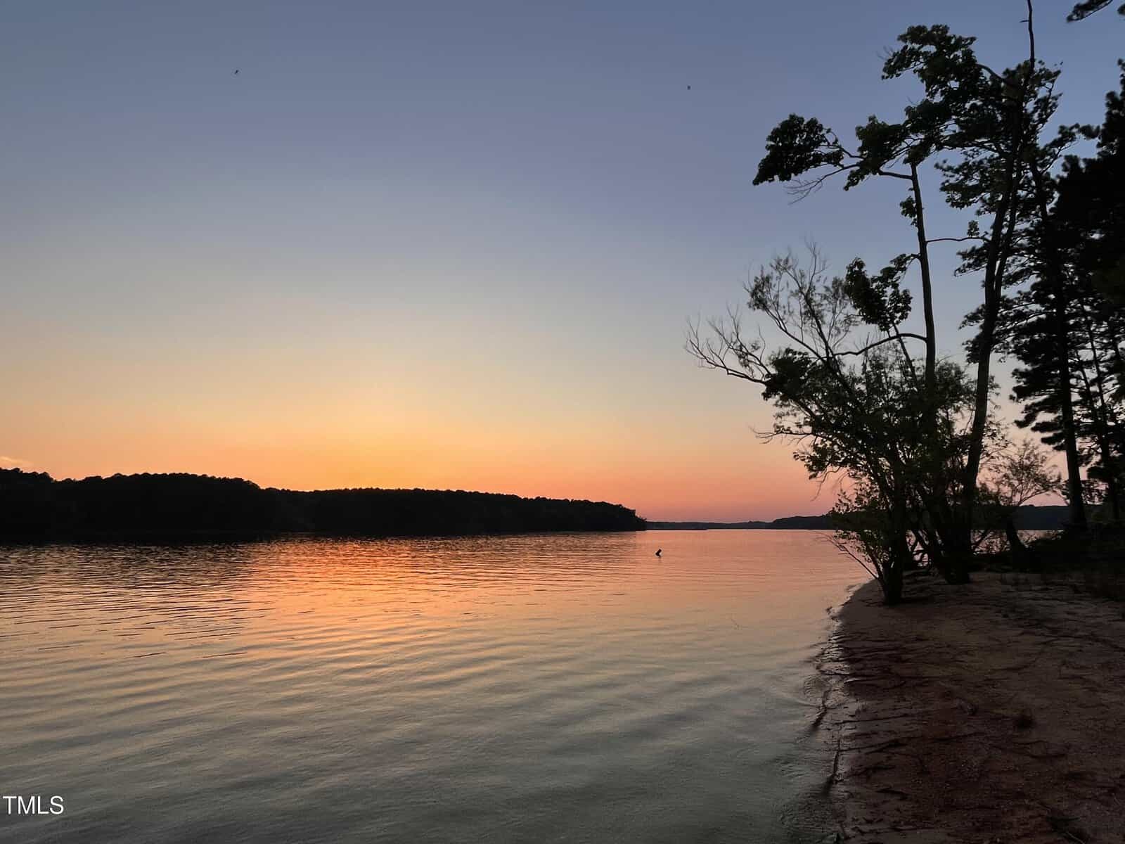 Sunset over a serene lake with calm waters gently reflecting the pastel hues of the sky. The silhouette of trees lines the right shoreline, while the dark outline of distant hills is visible against the fading light on the left horizon.