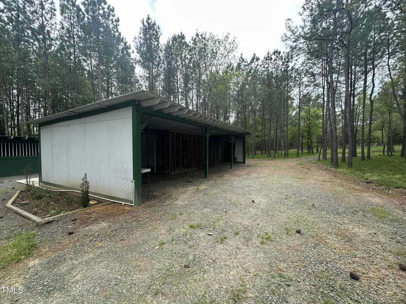 A large, open green shed with a metal roof stands on a gravel area. Surrounding the structure are tall pine trees and patches of grass, creating a rustic, forested setting under a cloudy sky.
