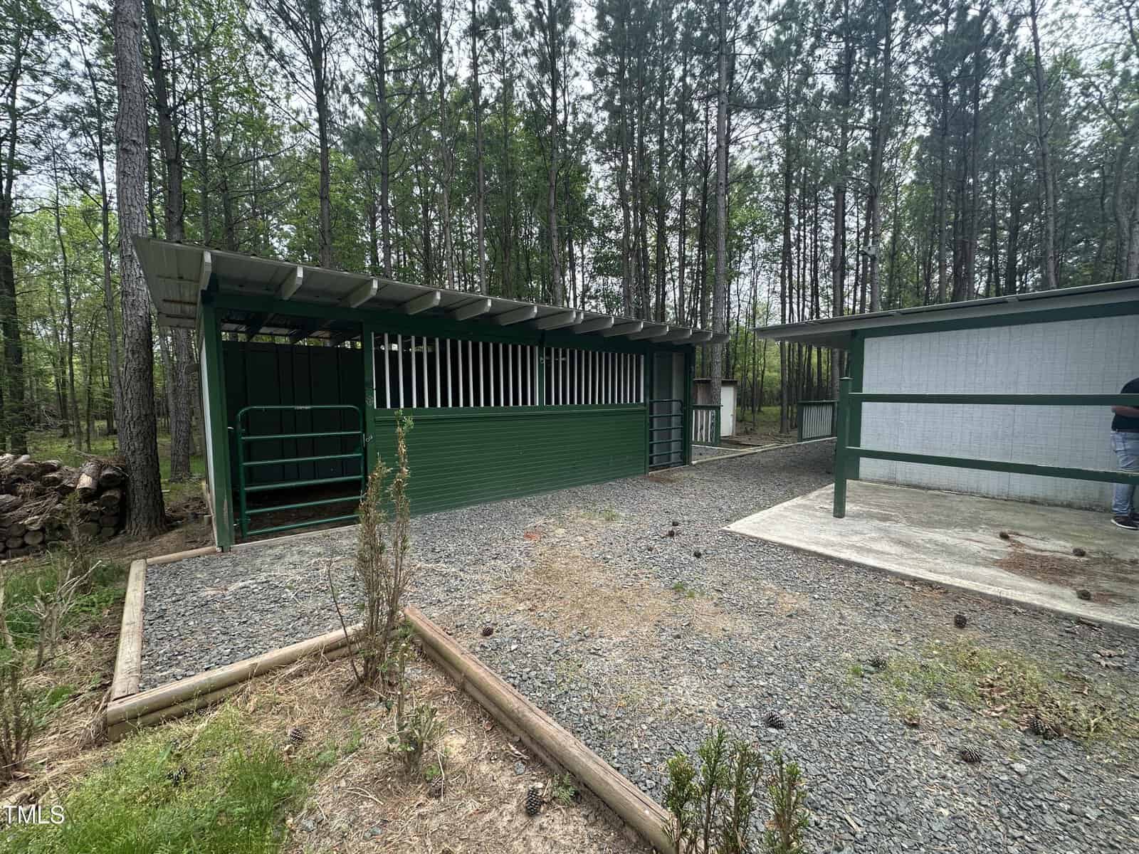 A gravel path leads to a green and white barn structure surrounded by tall trees. The barn features a covered area with vertical bars and corrugated metal roofing. Nearby, a person stands partially visible next to another structure.