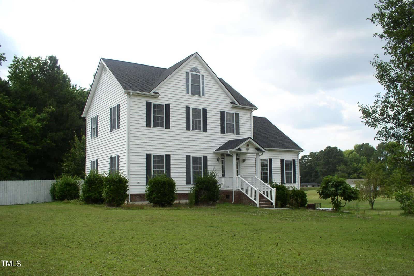 A charming two-story white house with black shutters and a triangular roof, set on a spacious green lawn at 90 Evergreen Place, Henderson, NC. It features a covered front porch with steps leading up, surrounded by neat shrubs. A white fence and trees are visible in the background under a partly cloudy sky.