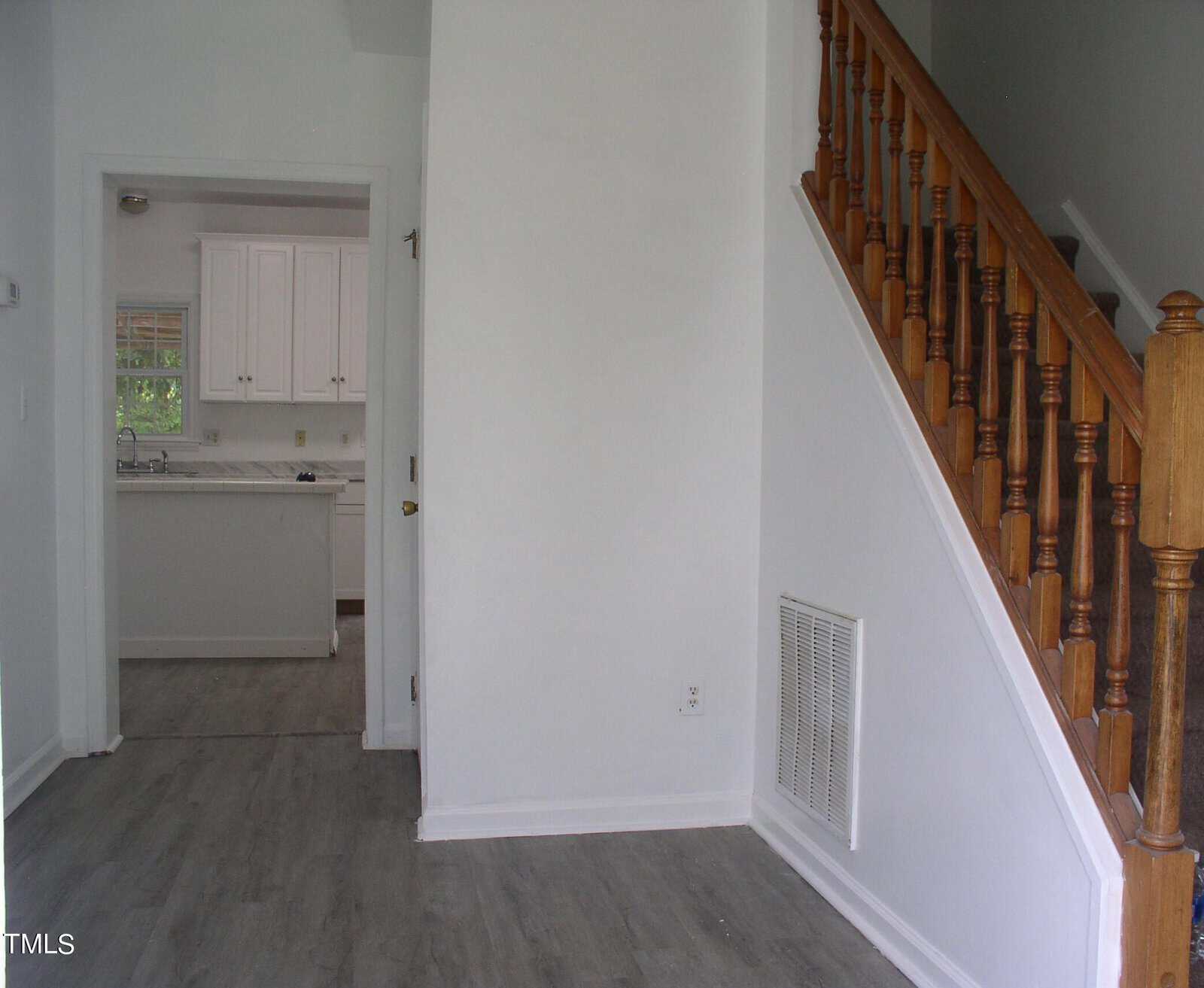 A small, empty living space is shown with light gray flooring and white walls. On the right, a wooden handrail and stairs lead upwards. An adjacent open doorway offers a view into a compact kitchen area featuring white cabinets and light-colored countertops.