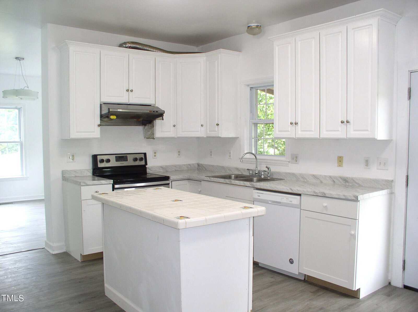A bright kitchen with white cabinets and a gray marbled countertop. It features an electric stove with a hood, a dishwasher, and a stainless steel sink beneath a window. The center island has a tiled top. The adjacent room is partially visible with ample natural light.