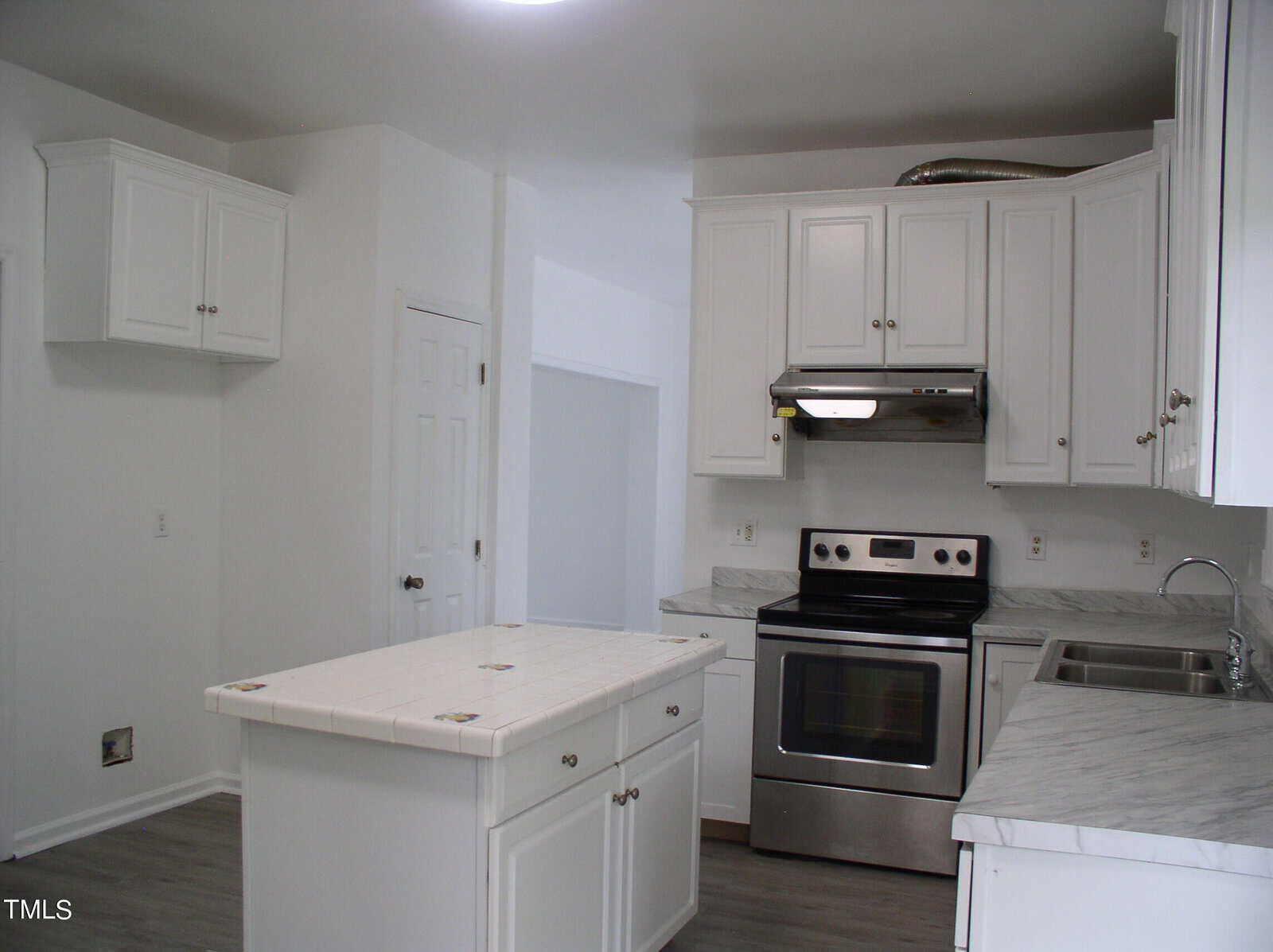 A modern kitchen with white cabinets, a white tile countertop island, stainless steel appliances including an oven and range hood, a marble-patterned countertop, gray flooring, and a double sink with a silver faucet under a white ceiling with built-in lights.