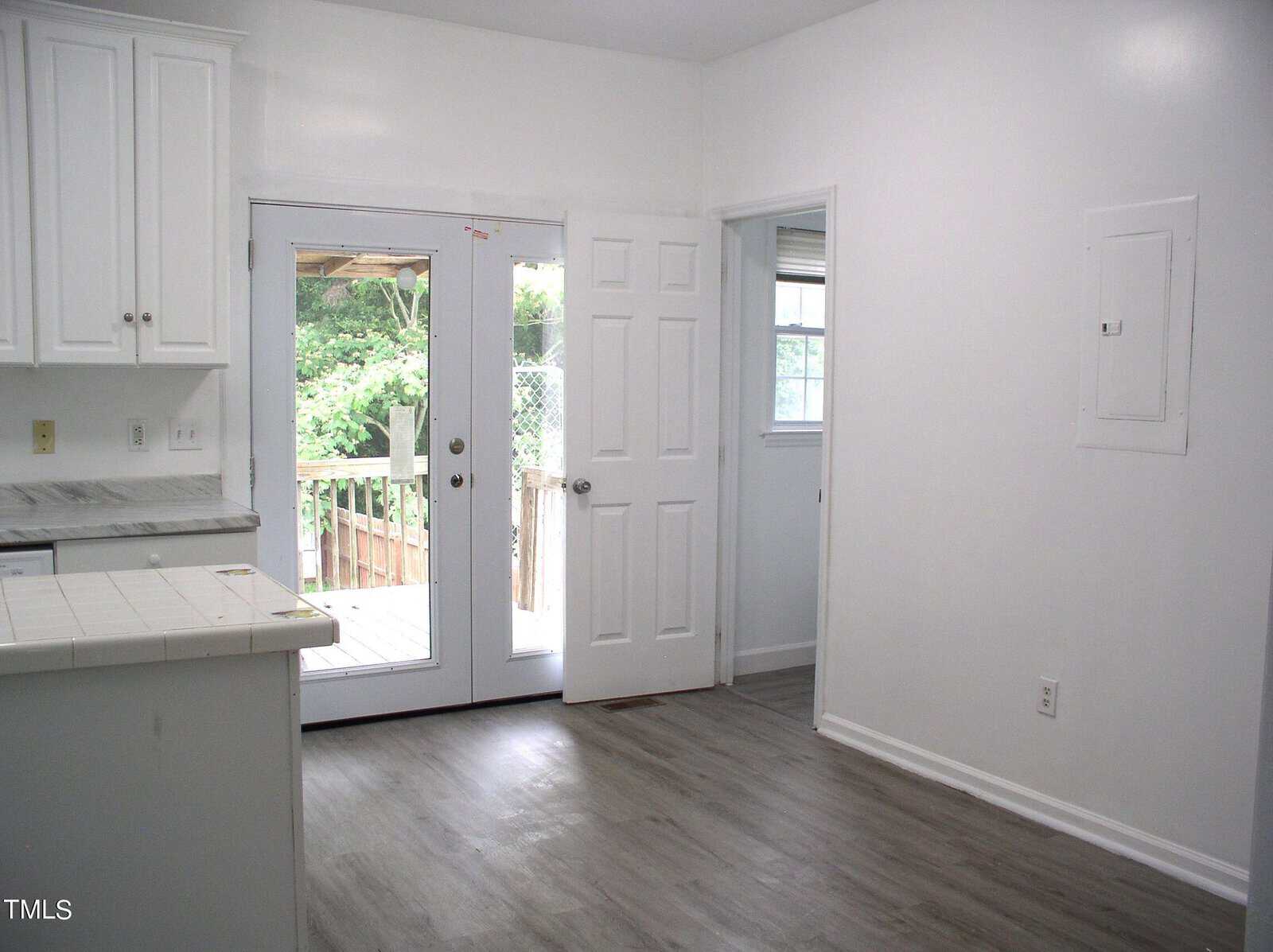 A bright kitchen with white cabinets, white tile countertops, and gray wood flooring. Double doors lead to an outdoor patio. An open doorway on the right reveals another room. The kitchen is simple and uncluttered, with natural light coming through the doors.