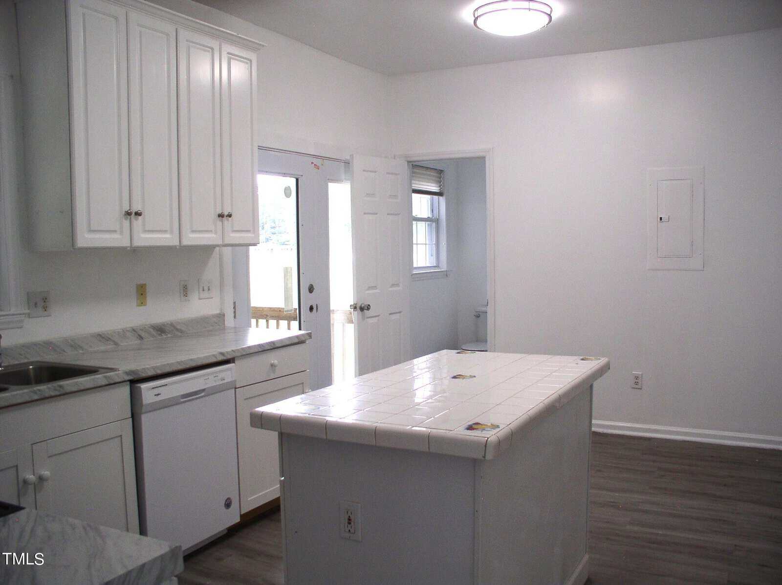 A modern kitchen with white cabinets, a white tiled countertop island, and stainless steel appliances, including a dishwasher and stove. A door with a window leads outside, and a window above the sink lets in natural light. The room has gray hardwood flooring.