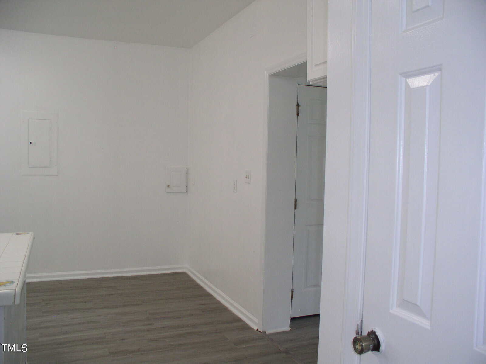 A plain white room with light gray wooden flooring. The room has a doorway on the right leading to another room and a small access panel on the left wall. The walls and door are white, giving the space a minimalist appearance.