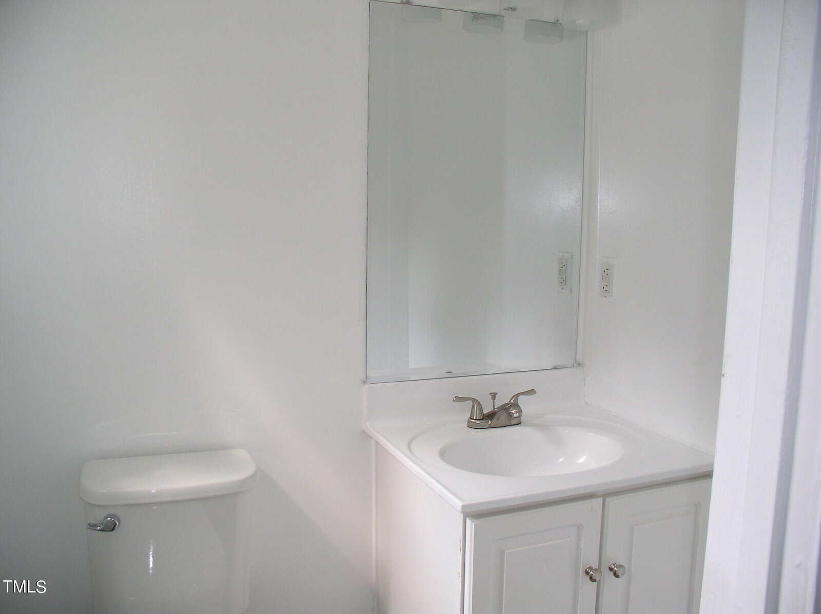 A small bathroom with white walls features a toilet on the left and a white vanity with a sink and faucet on the right. Above the sink, a rectangular mirror is mounted on the wall. Two electrical outlets are visible to the left of the mirror.