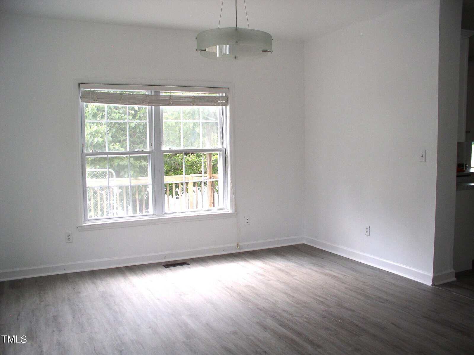 A vacant room with white walls and light wood flooring. A window with blinds partially raised lets in light, illuminating the space. A modern circular light fixture hangs from the ceiling. Part of a kitchen area is visible to the right.