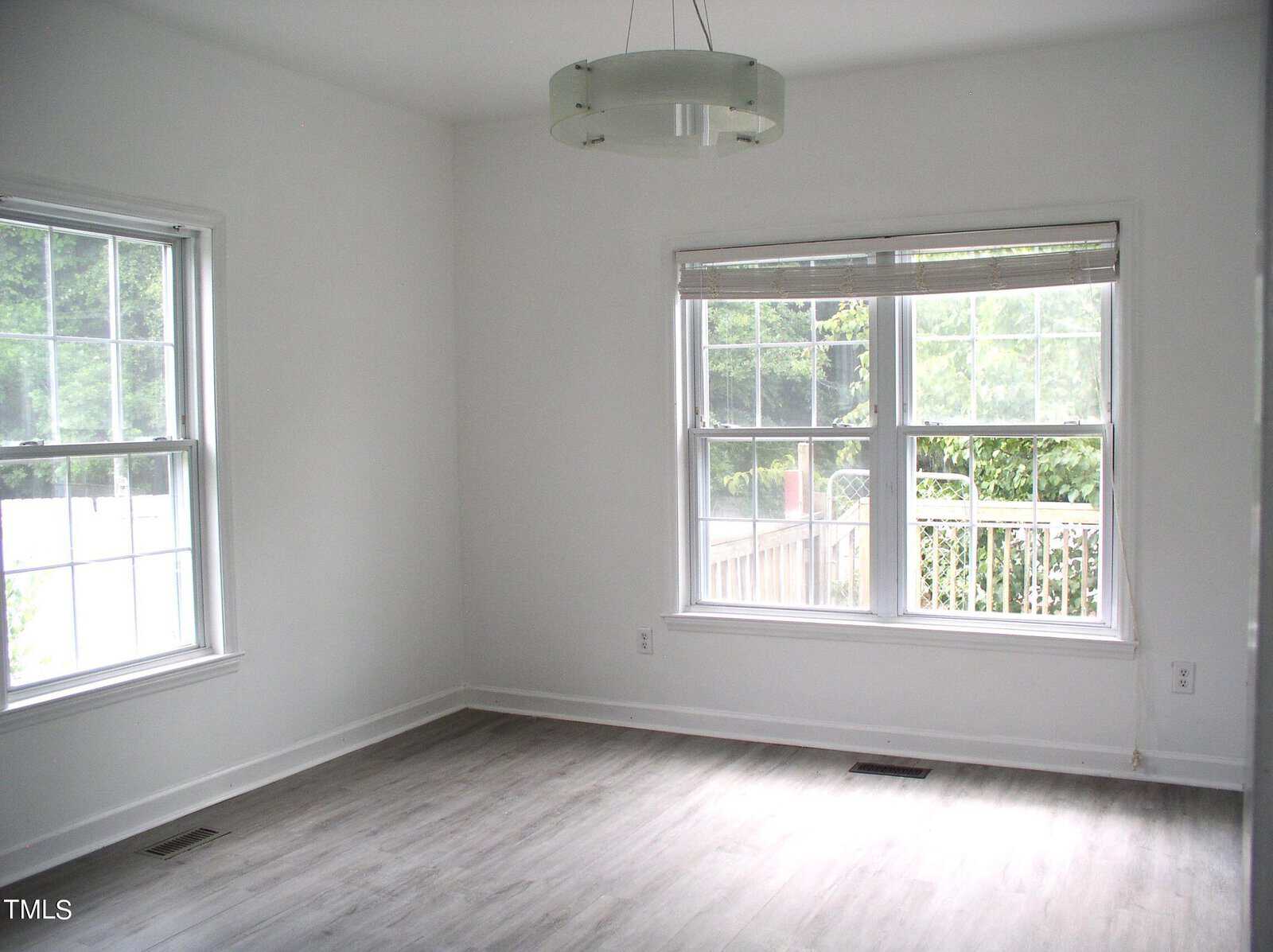 A bright, empty room with light gray wood flooring, white walls, and two large windows letting in natural light. A modern circular light fixture hangs from the ceiling. Outside, greenery is visible through the windows.
