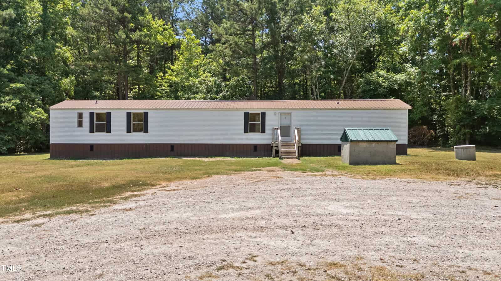 A white mobile home with a brown base sits on a spacious, grassy lot at 274 Brookston Road in Henderson, NC, surrounded by tall trees. The home features a few small windows and a green metal roof over a nearby structure. A dirt driveway leads up to the front steps and door of the mobile home.