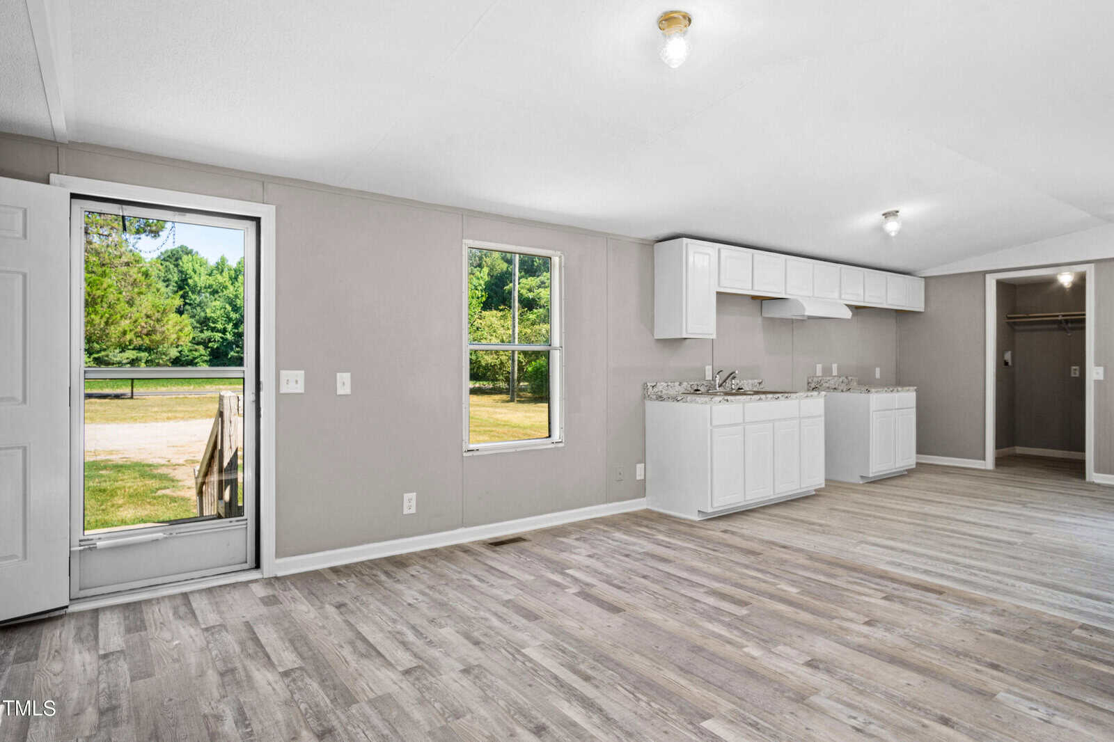 A spacious, light-filled kitchen with white cabinets and countertops. The room features light wood flooring and a large window overlooking a green yard. A door to the left opens to the outside, bringing in more natural light. The walls are painted in a soft gray tone.