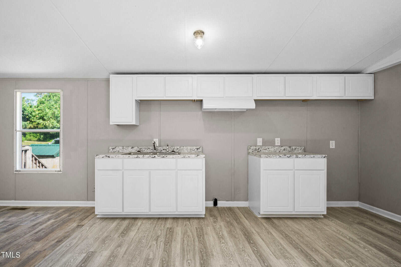 A spacious kitchen with light gray walls and wood-patterned vinyl flooring. It features white upper and lower cabinets with marble countertops. A window is on the left wall, providing natural light, and a single light fixture hangs from the ceiling.