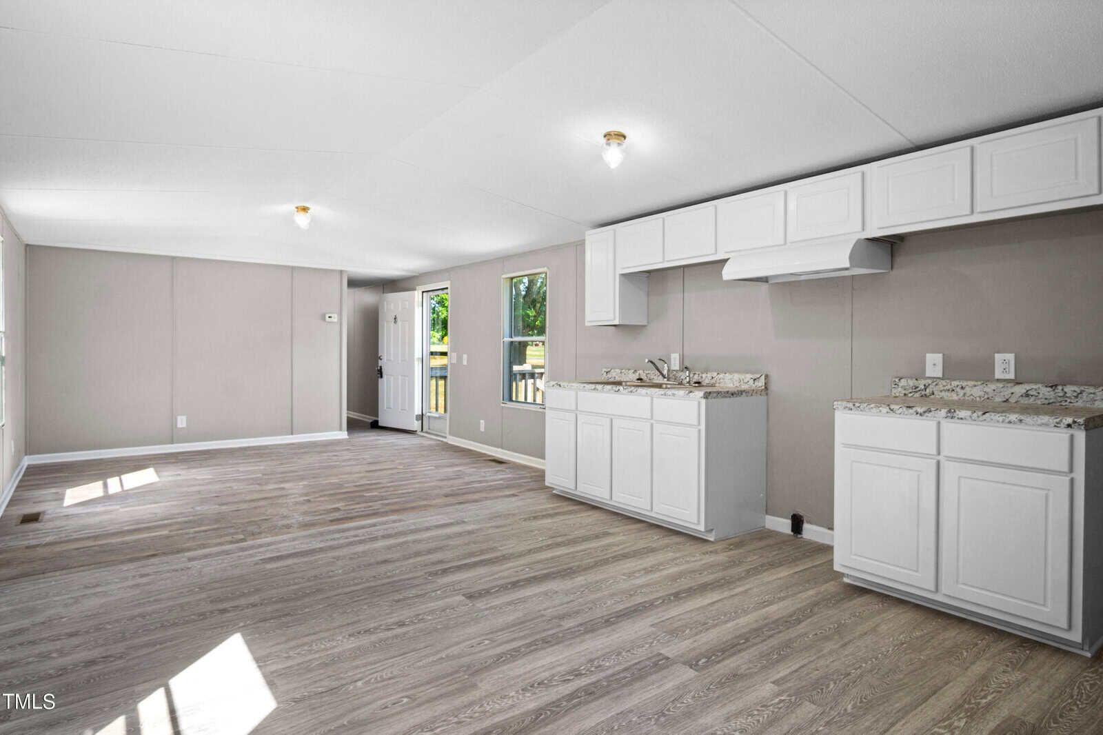A spacious, empty kitchen and living area with light gray walls and wood flooring. The kitchen has white cabinets, a marble countertop, and a vent hood. Two windows and a doorway provide ample natural light, along with recessed ceiling lights. A door leads outside.