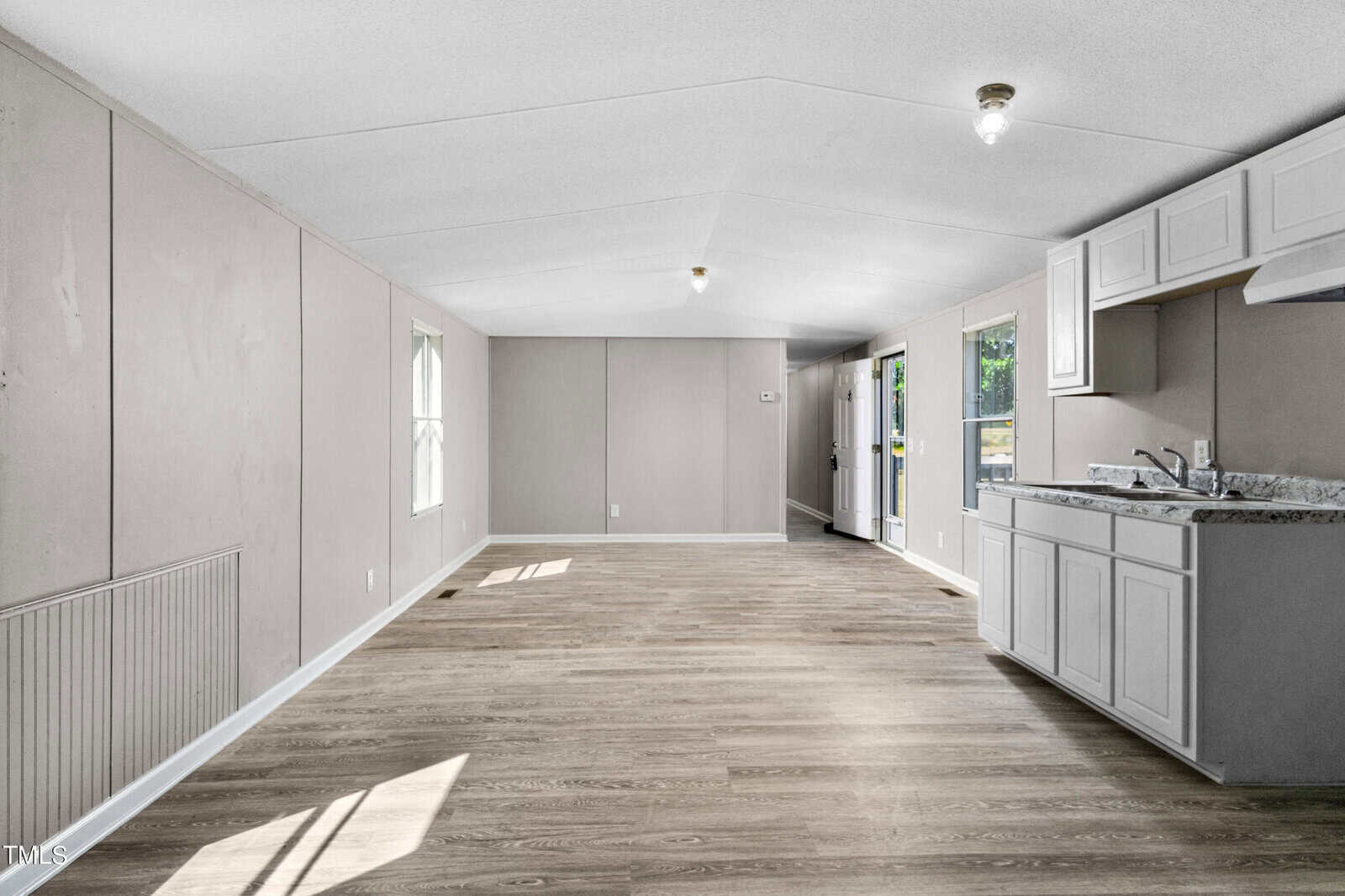 A brightly lit, unfurnished room with light grey walls and wooden flooring. The room has two windows on the left side, a small light fixture on the ceiling, and a kitchen area with white cabinets and a sink to the right. A door and additional windows are at the far end.