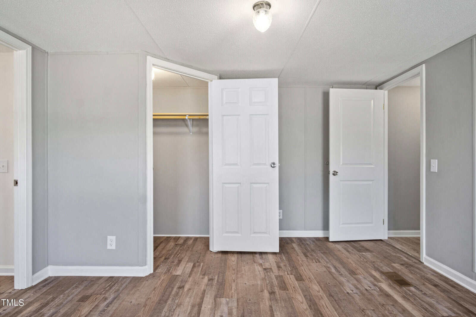 A modern, empty room featuring light gray walls and a ceiling light. The room has wooden flooring and two white doors - one leading to a walk-in closet and the other to an adjacent space. A light switch and electrical outlet are visible on the left wall.