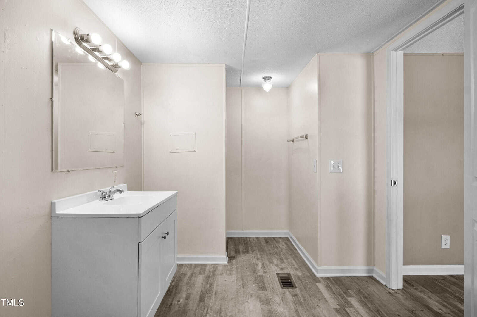 A bathroom with a white vanity and sink under a lighted mirror on the left. The room has beige walls, a wooden floor, and a light fixture on the ceiling. A towel rack is mounted on the far wall, and an open doorway is on the right.