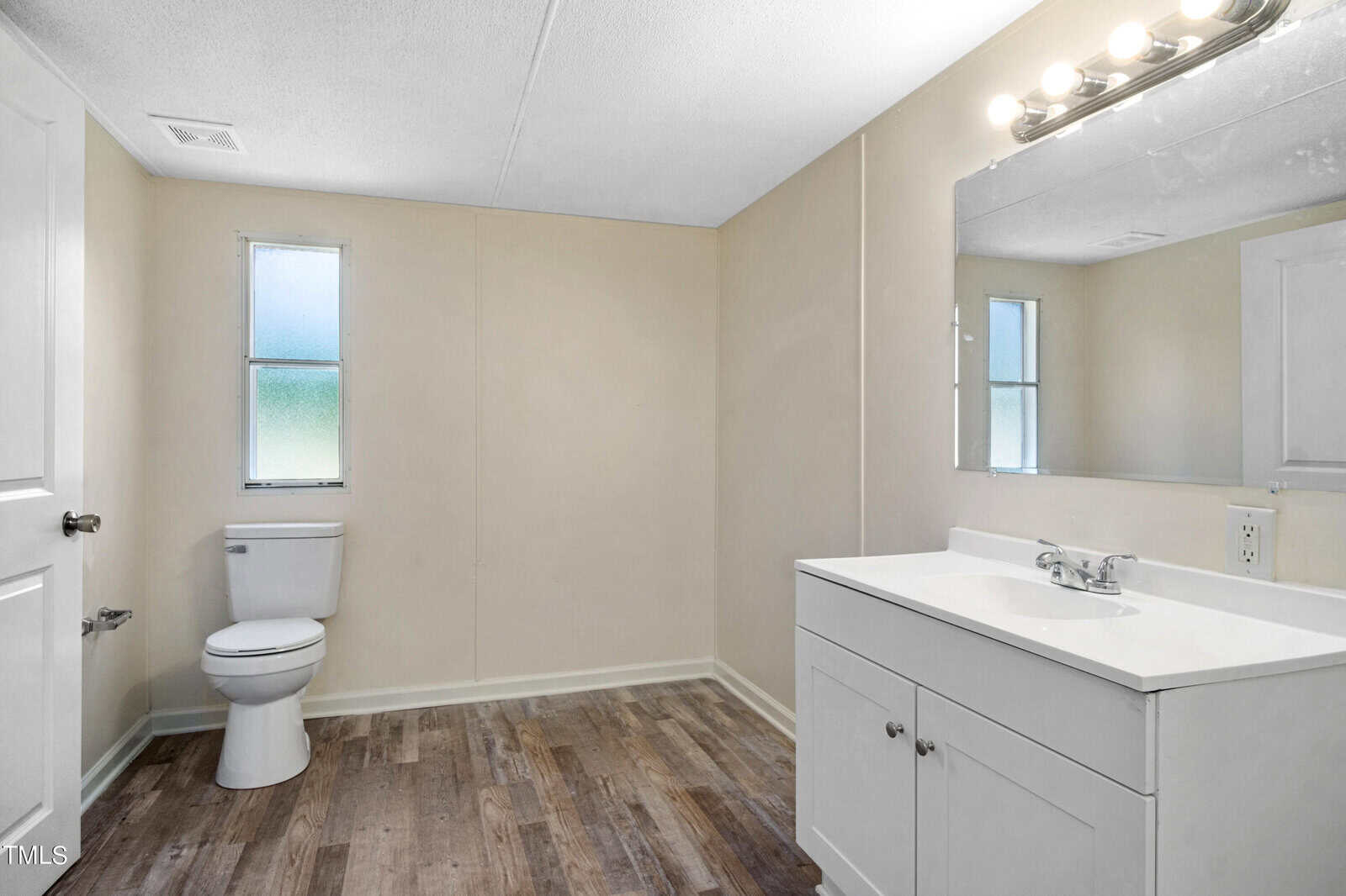 A bright, minimalistic bathroom featuring a white toilet and vanity with a sink, faucet, and large mirror. Two frosted windows allow natural light. The floor is made of wood-look vinyl, and the walls are painted a light beige color. A row of lights is above the mirror.