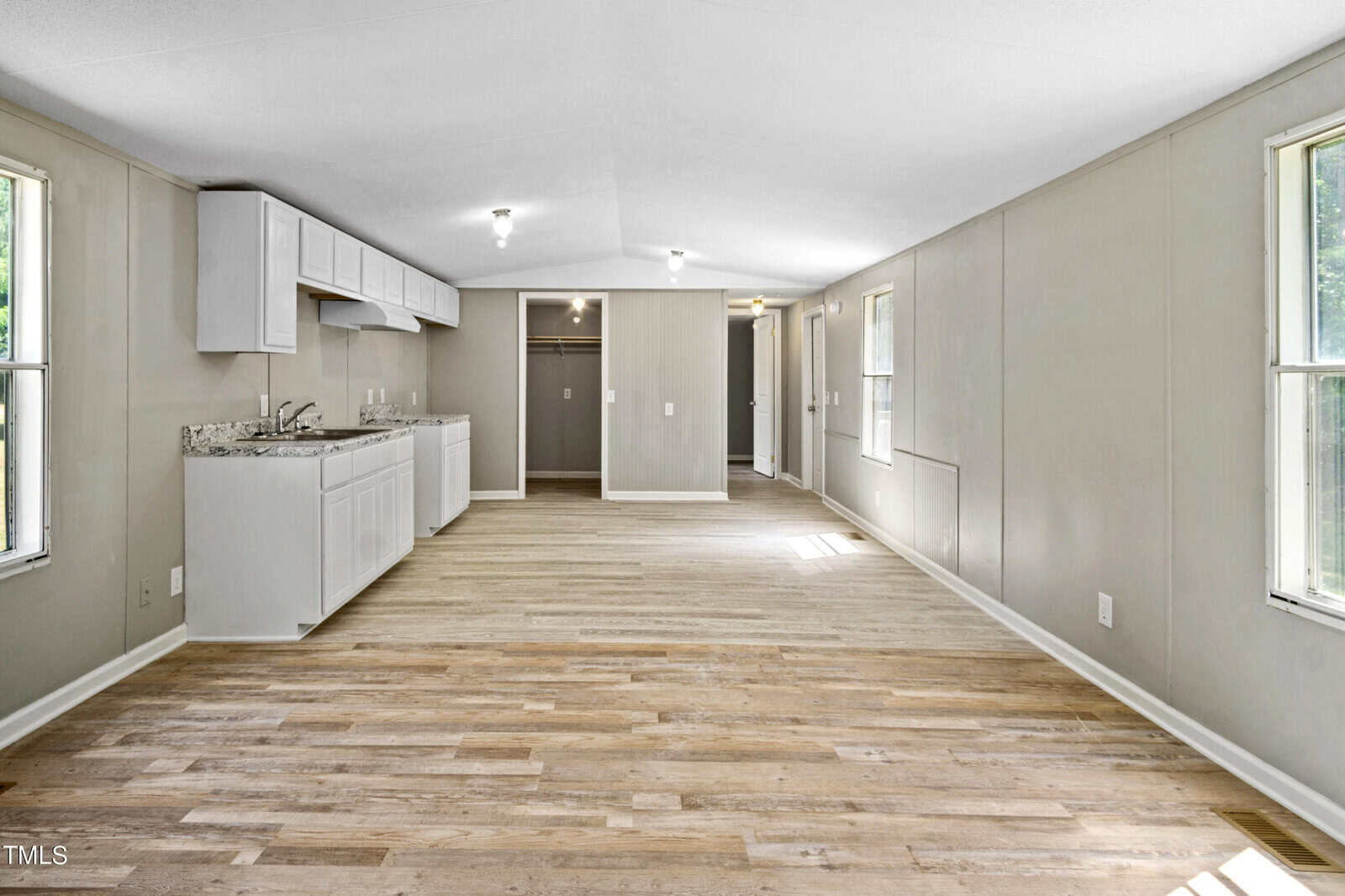 A spacious, empty room with light wooden flooring and beige walls. At one end, the room features a kitchen area with white cabinets and a granite countertop. Two large windows are on the right wall, allowing natural light to fill the space.