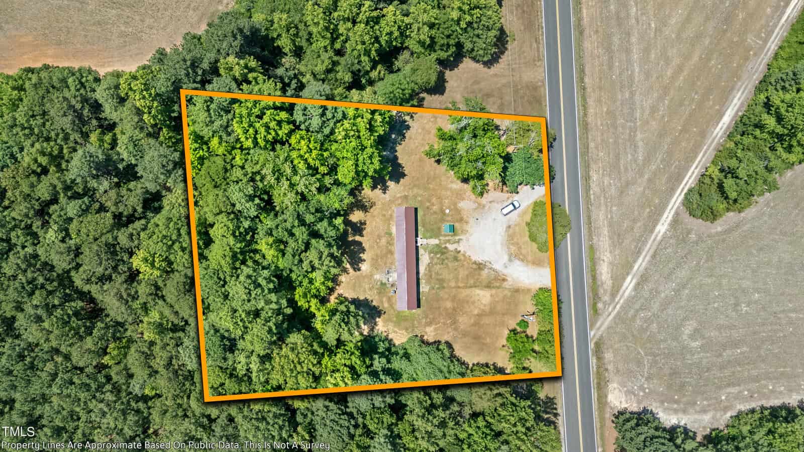 Aerial view of a rectangular plot of land outlined in orange, located adjacent to a main road. The plot contains a long, narrow building surrounded by trees and open areas. The building is situated closer to the center and left side of the property.