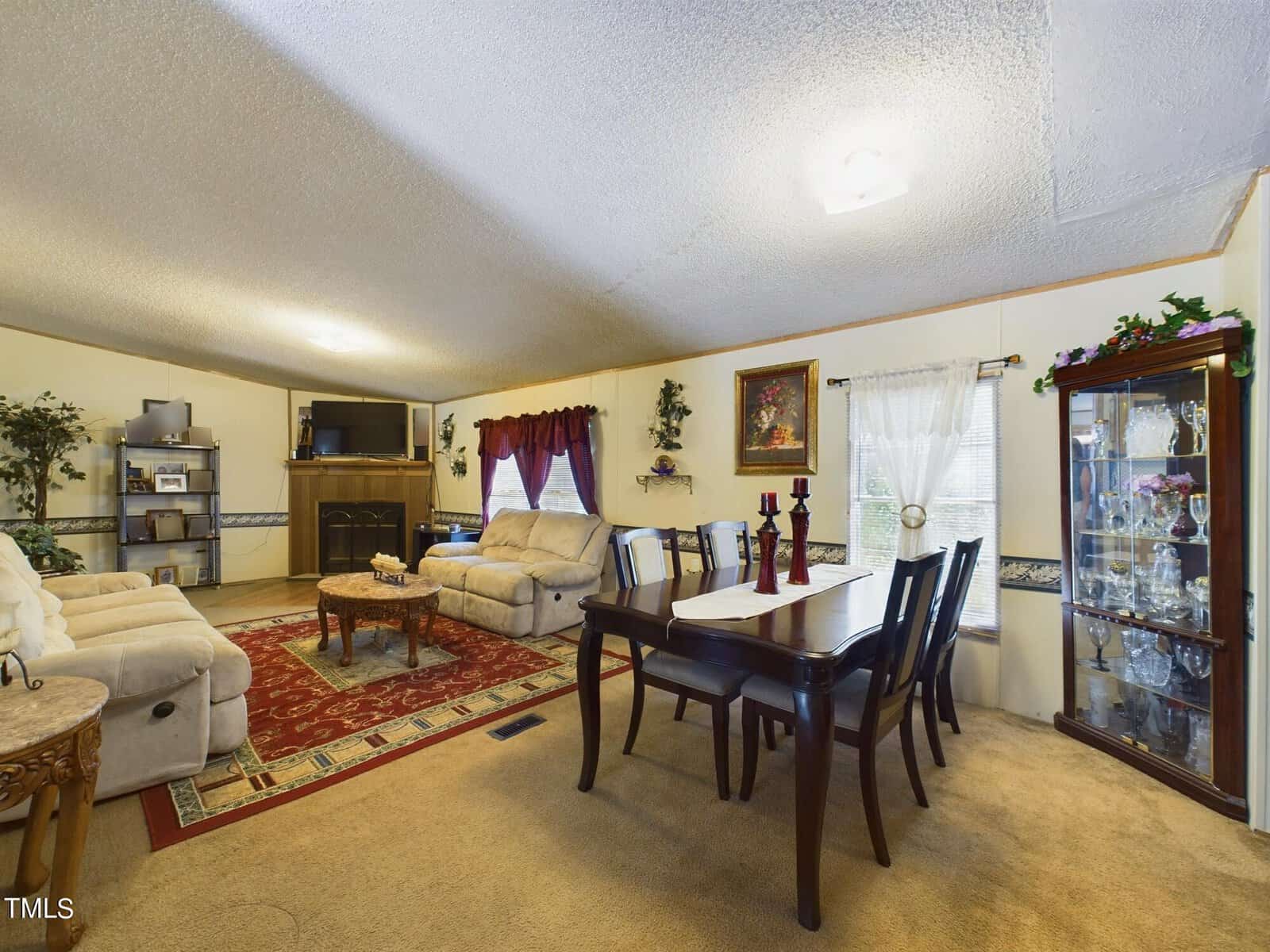 A cozy living room with a dining area. Beige sofas face a TV and fireplace. A red patterned rug covers the floor. A dining table with chairs is on the right, and a glass cabinet filled with chinaware stands nearby. Wall decorations add warmth.