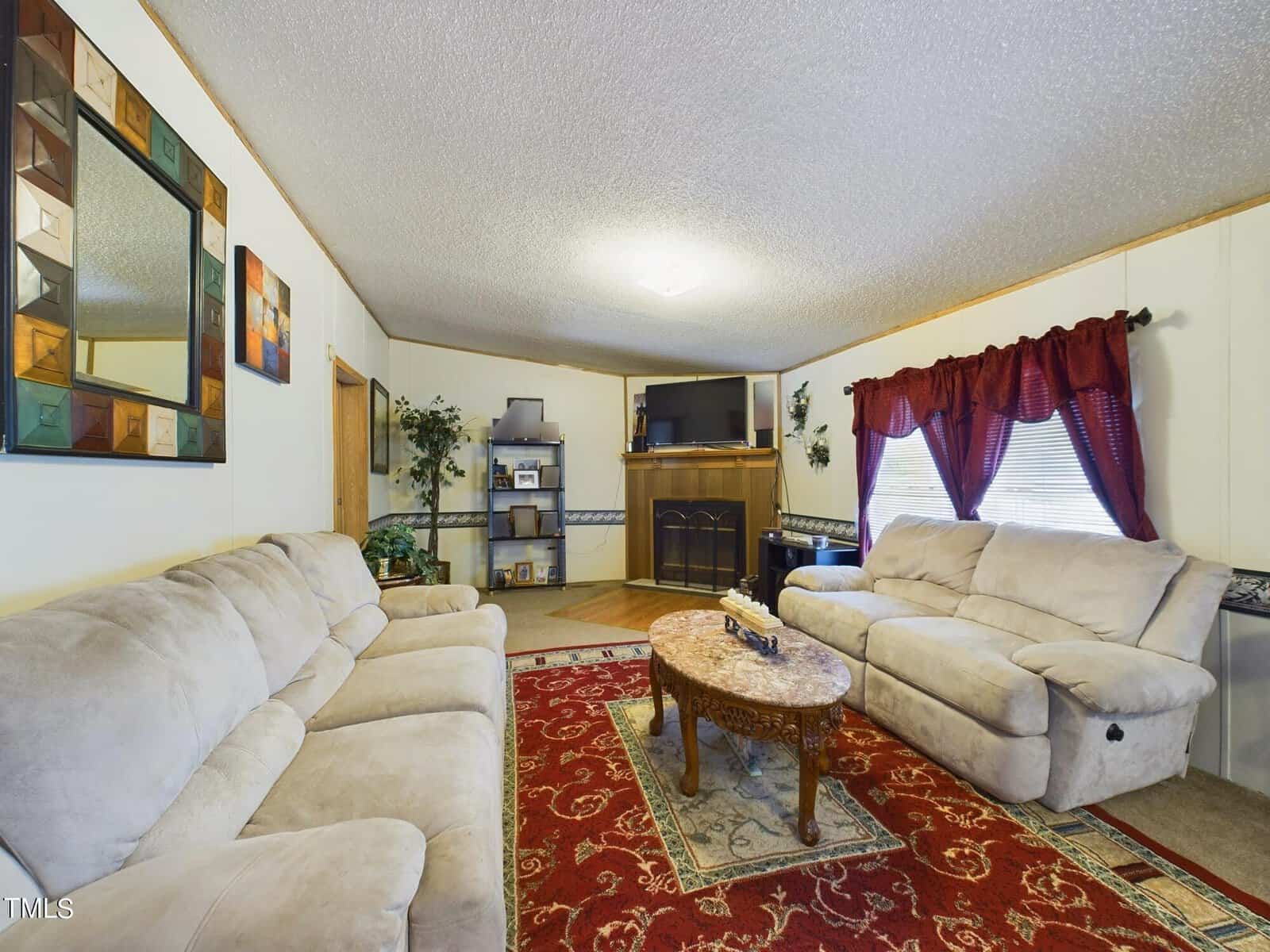 Cozy living room with two beige sofas, a red patterned rug, and a wooden coffee table. A flat-screen TV sits on a shelf above a fireplace. Large windows with red curtains bring in natural light. Decorative plants and a wall mirror complete the decor.