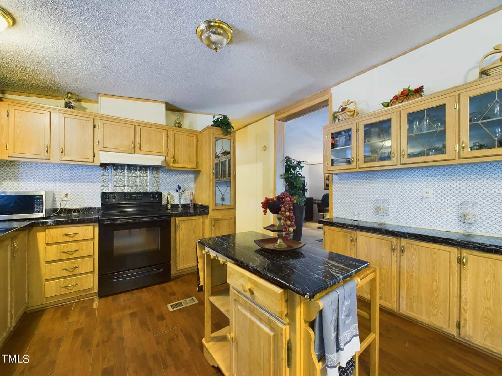 A kitchen with wooden cabinetry, black countertops, and a black stove. A small island with a vase of flowers is in the center. Glass cabinets hold dishes. The room has a warm, rustic feel.