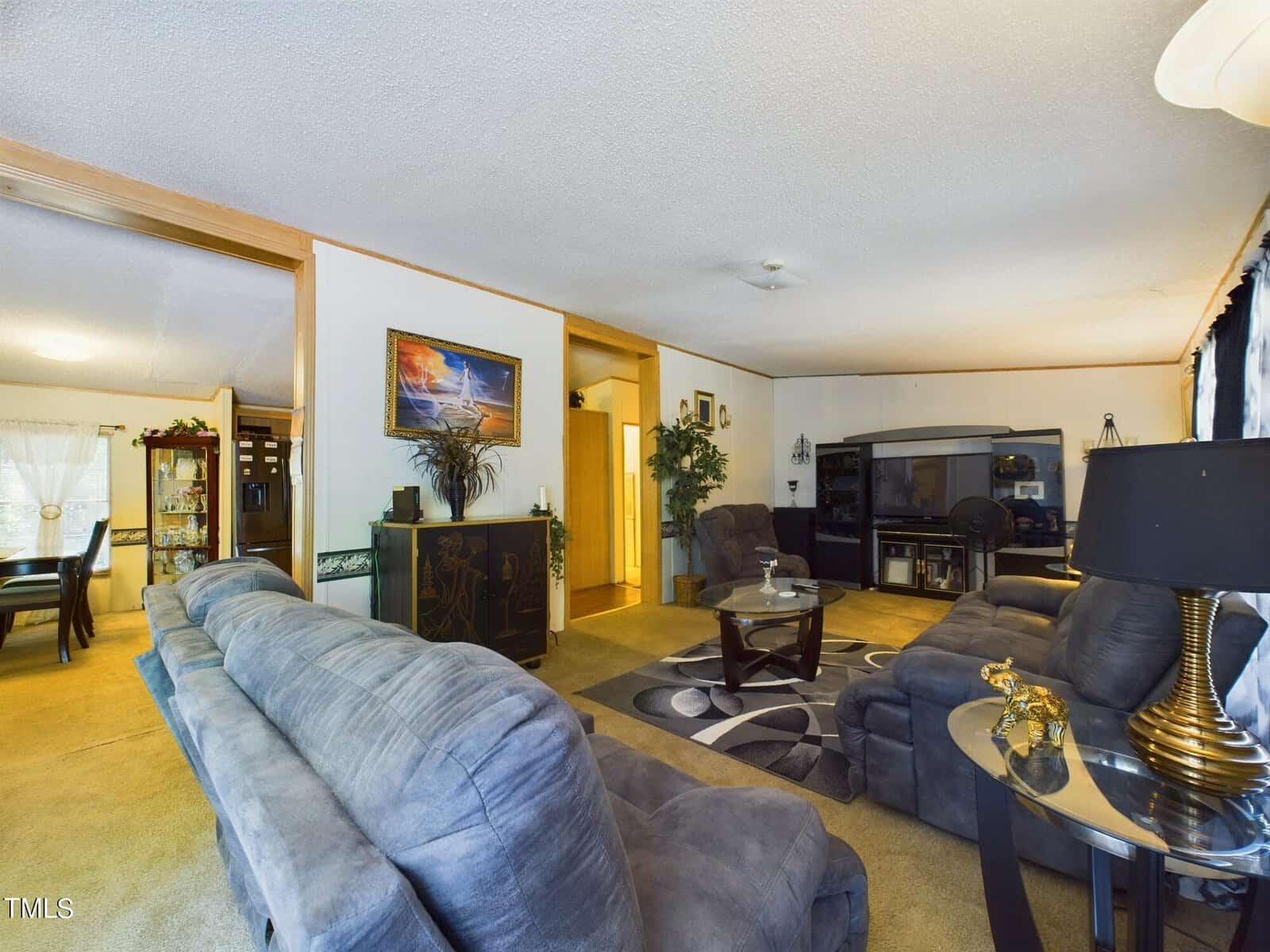 A cozy living room with a blue sofa and armchairs, a round glass coffee table, and a large TV in the corner. The room has carpeted floors, decorative plants, and a dining area visible in the background through an open doorway.
