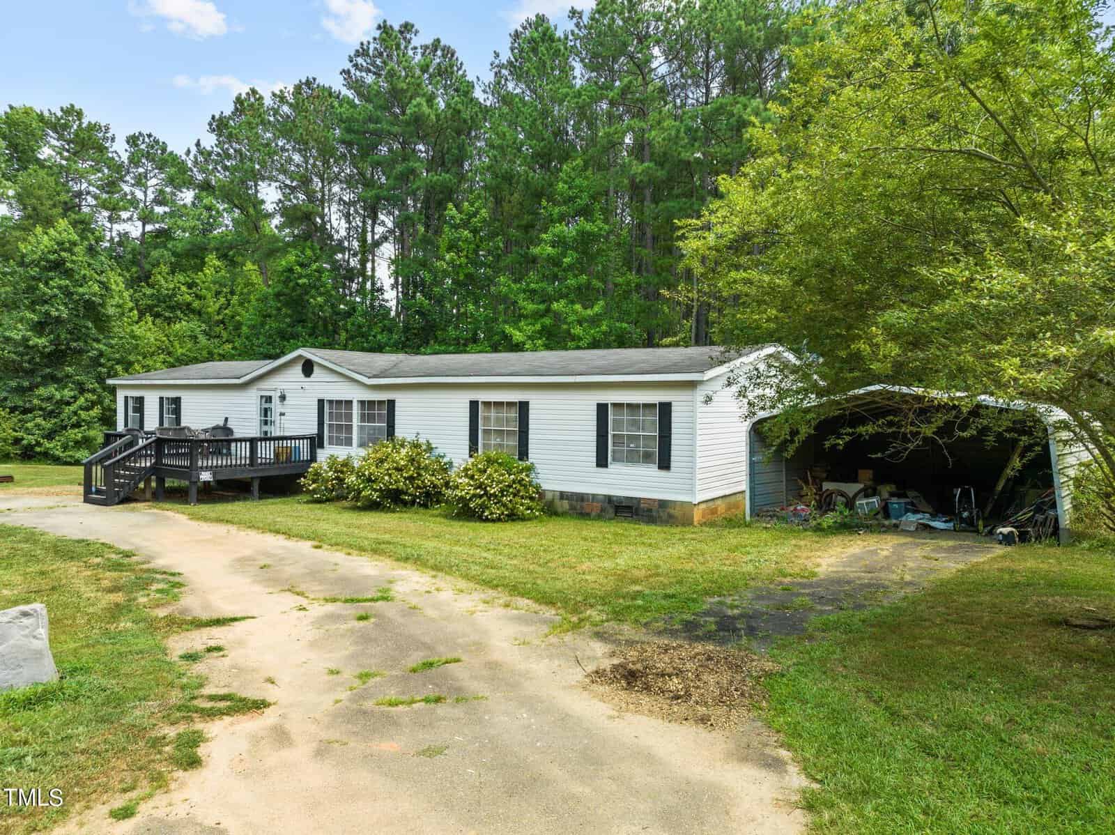 Located at 1144 Nottingham Way in Creedmoor, this white manufactured home boasts a lush green front yard with trees as a scenic backdrop. A small wooden deck complements the attached carport, and the curved driveway adds charm to the inviting property.