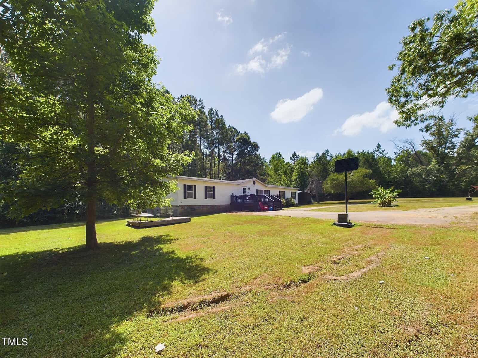 A white house with a front porch surrounded by trees and a spacious lawn on a sunny day. A gravel driveway leads to the house, which is set against a backdrop of tall trees and a clear blue sky.