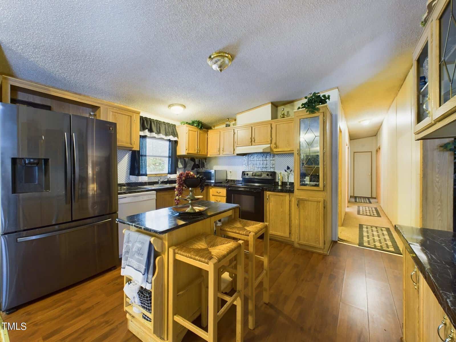 A modern kitchen with wooden flooring and cabinetry features a large stainless steel refrigerator, an island with bar stools, and a built-in oven. The countertops are dark, and there is a hallway leading to other rooms.