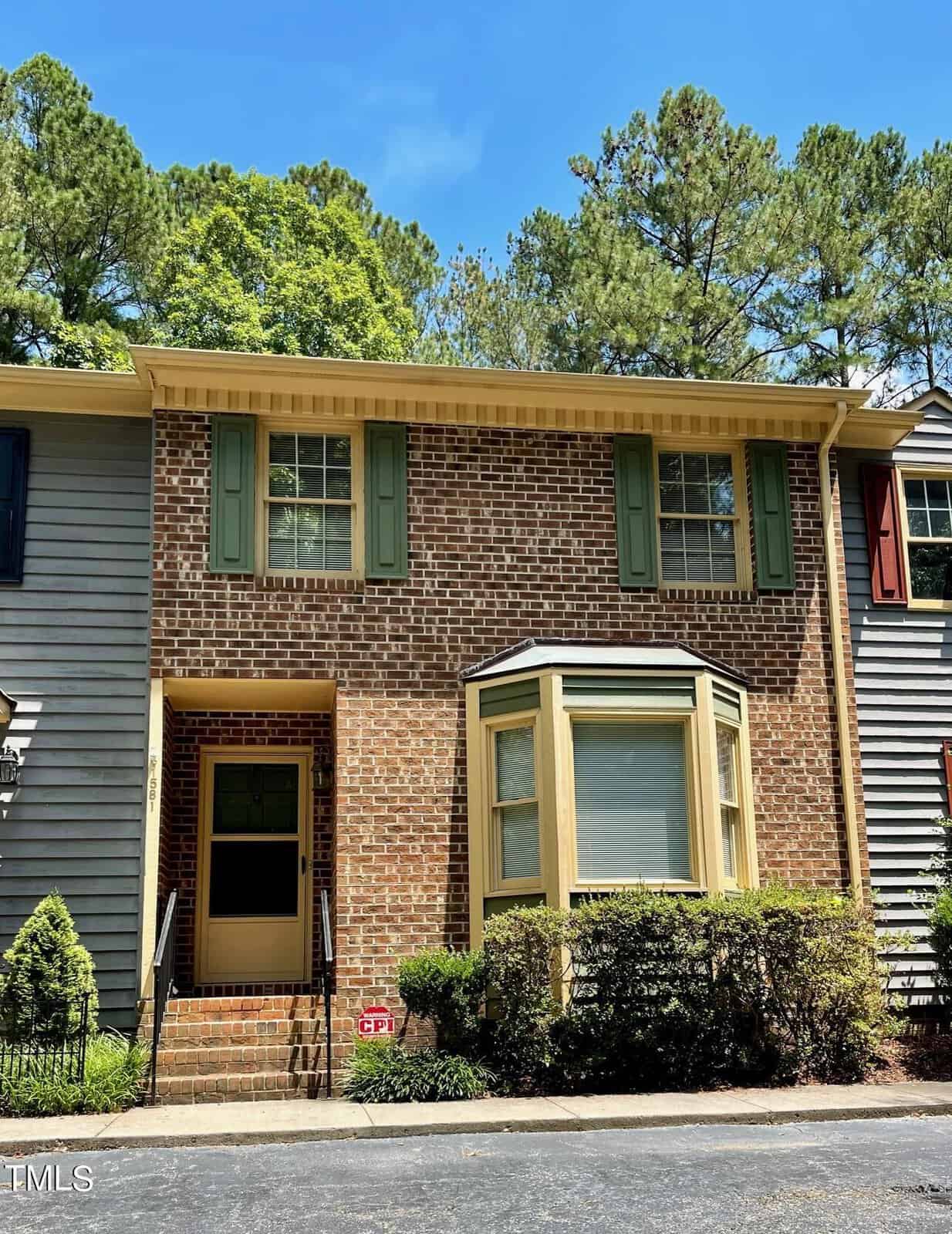 A charming two-story brick townhouse at 1581 Graham Avenue Unit 3 in Henderson, NC, features green shutters and a bay window on the first floor. The front entrance boasts black railing and steps leading to a wooden door with a glass panel. The surrounding area is adorned with shrubs and trees under a clear blue sky.