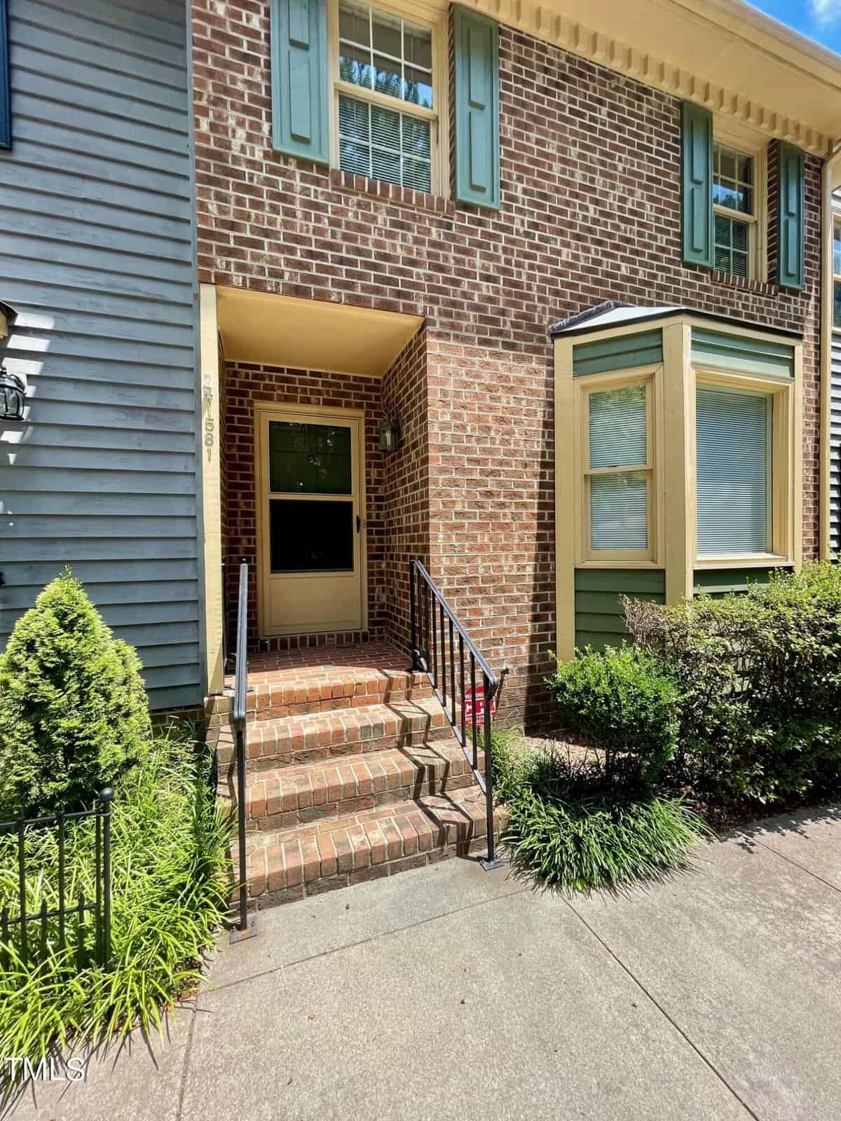 A brick townhouse with a green bay window and shutters. The front door is accessed by a short flight of brick steps with a black metal railing. There are green plants lining the walkway leading to the door. The house number "1689" is visible.