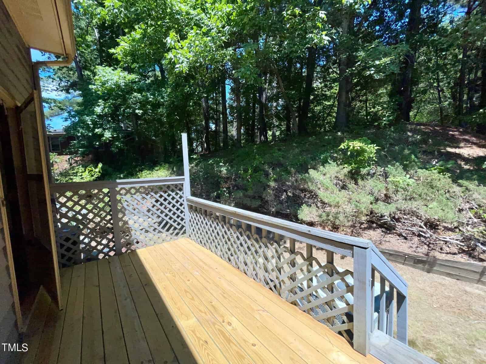 A wooden deck attached to a house overlooks a wooded area. The deck features lattice railing at the side and open view toward the trees. The scene is bathed in sunlight, highlighting the natural surroundings.
