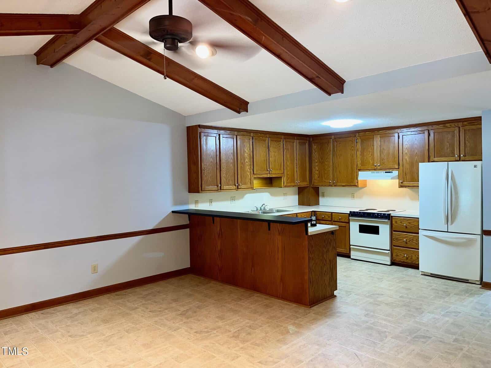 A kitchen with wooden cabinets and a white refrigerator, stove, and hood. It features a light-colored tile floor, a ceiling fan with lights, and exposed wooden beams on the ceiling. A counter with a sink and bar seating separates the kitchen from the adjacent area.