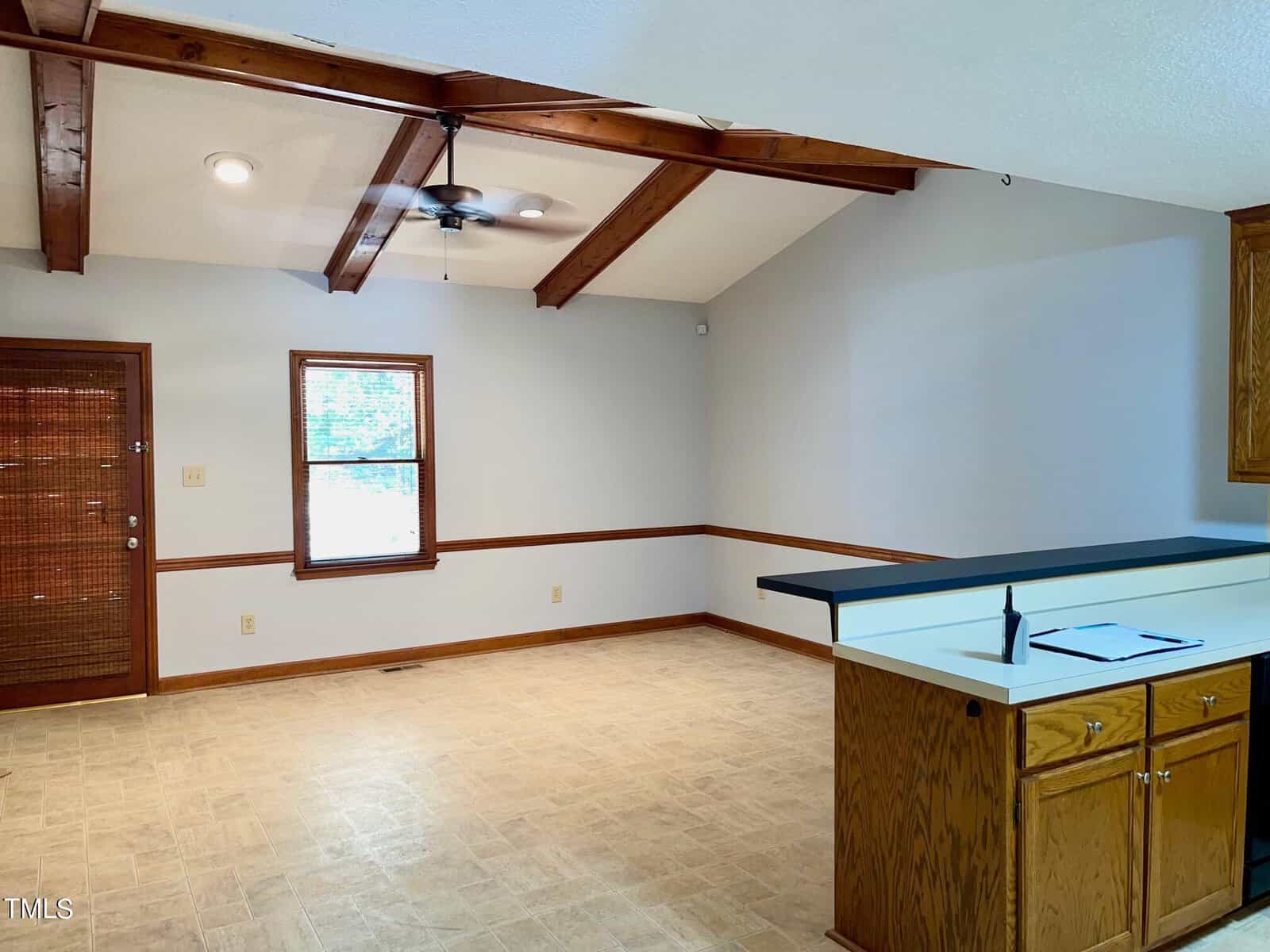A room with high ceilings and wooden beams, featuring a window, a door with wooden accents, and neutral-colored walls. There is a counter with a sink and cabinets on the right side. The flooring is light-colored, and the room appears well-lit by natural light.