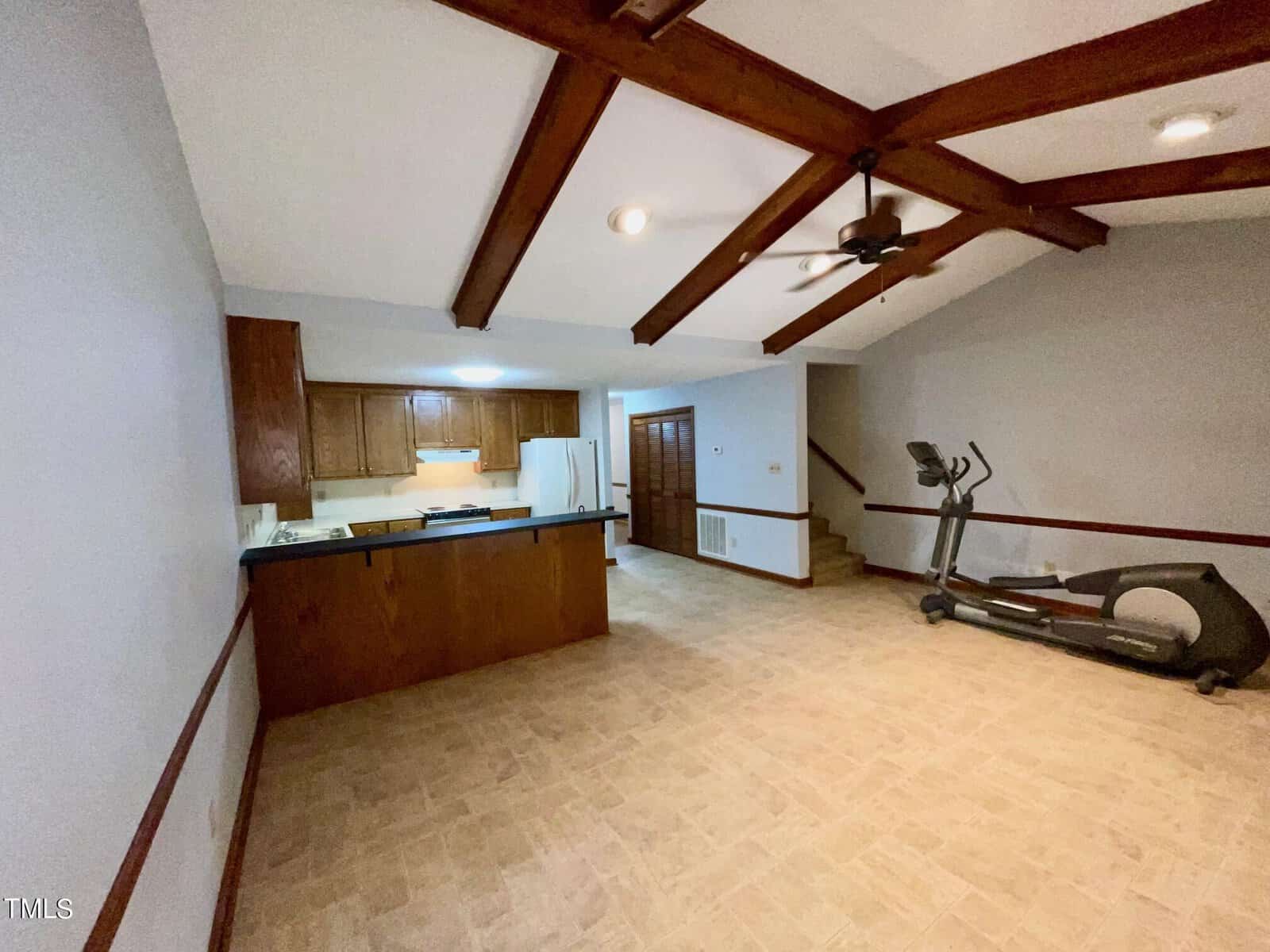 The image shows a spacious room with light-colored tile flooring, exposed wooden ceiling beams, and a ceiling fan. The room features a small kitchen area with wooden cabinets and white appliances, as well as an elliptical machine near the stairway.