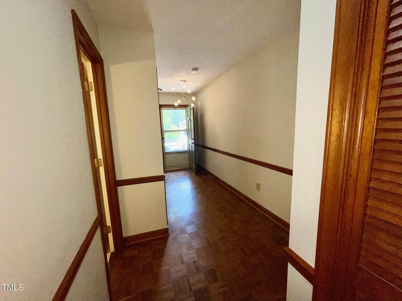 A hallway with wooden paneling on the lower half of the walls and wooden floor. The hallway leads to a door and window at the far end. There are doors on the left side and a louvered door on the right. Ceiling lights illuminate the space.