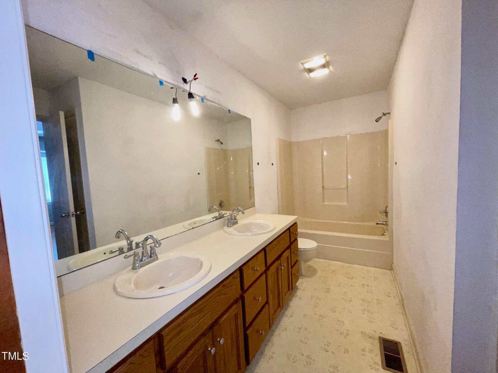 A bathroom with a double sink vanity, large mirror, wood cabinets, beige bathtub with shower, and light fixture on the ceiling. The floor has a light-colored pattern.