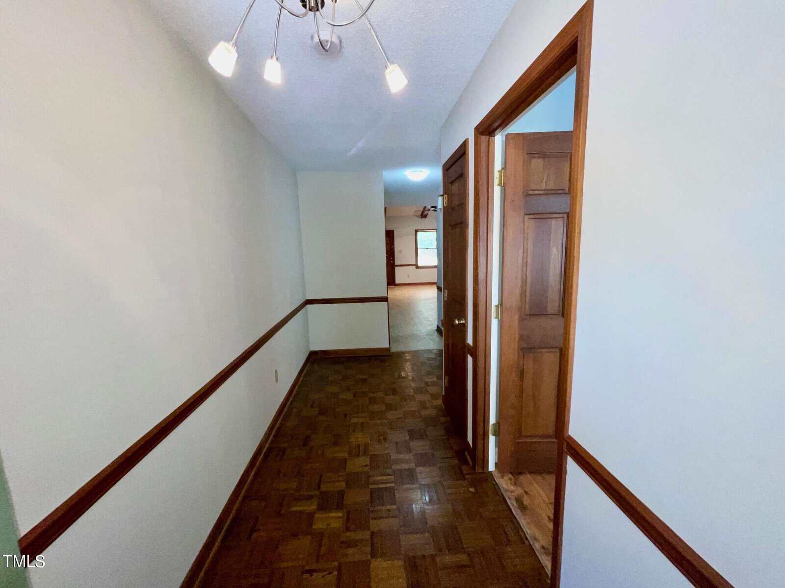 A hallway with parquet flooring, white walls with dark wood trim, and a light fixture on the ceiling. The hallway leads to a larger room in the background, with wooden doors on the right side and partially visible light fixtures.