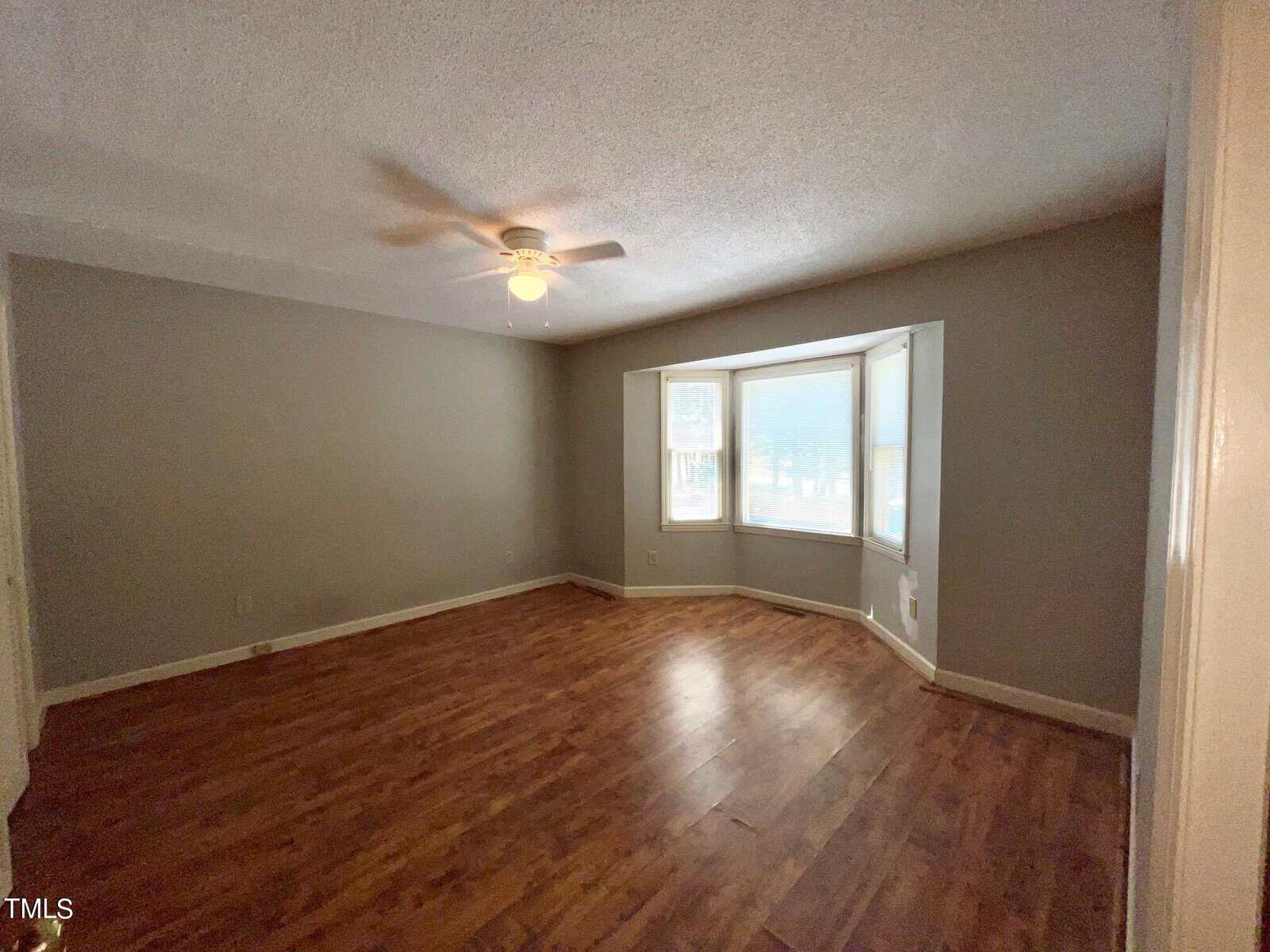An empty room with light wood flooring, gray walls, a ceiling fan, and a bay window with three sections allowing natural light to fill the space. The corner of an open door is visible in the foreground.