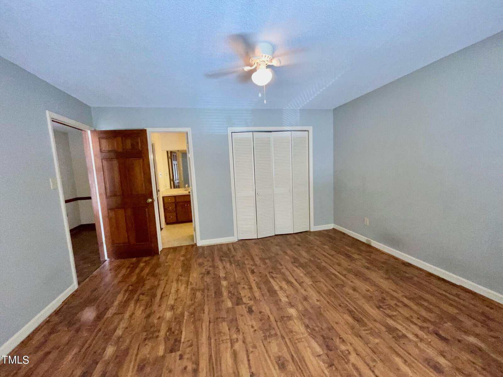 A room with light blue walls and wood flooring features a ceiling fan with a light. There is a closet with white louvered doors and an open door leading to a bathroom with visible sink and mirror. Another door is partially open to the hallway.