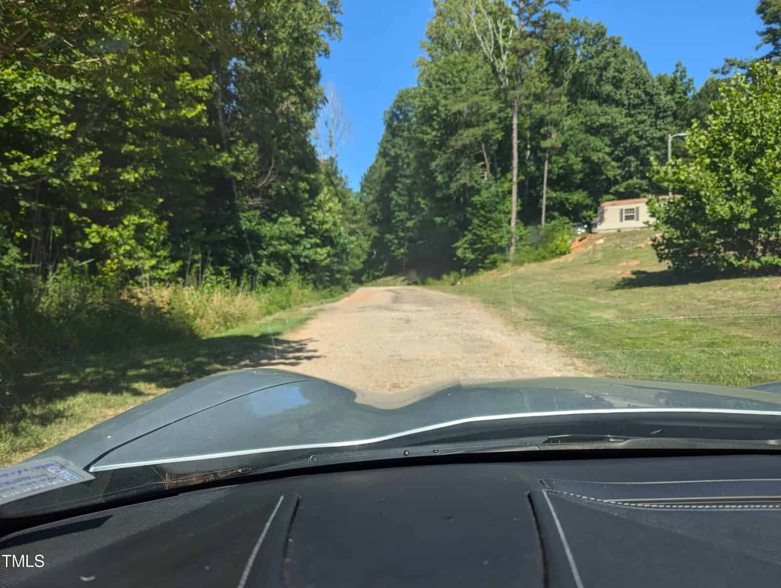 View from a car driving on a dirt road surrounded by trees. A small house is visible on the right side of the road. The scene is brightly lit by sunlight.