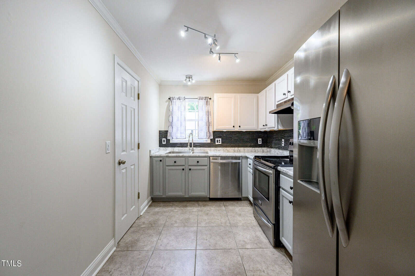 A modern kitchen with stainless steel appliances, including a fridge and oven. The cabinets are white and gray, and there's a window with sheer curtains. The floor is tiled, and the lighting is bright with ceiling spotlights.