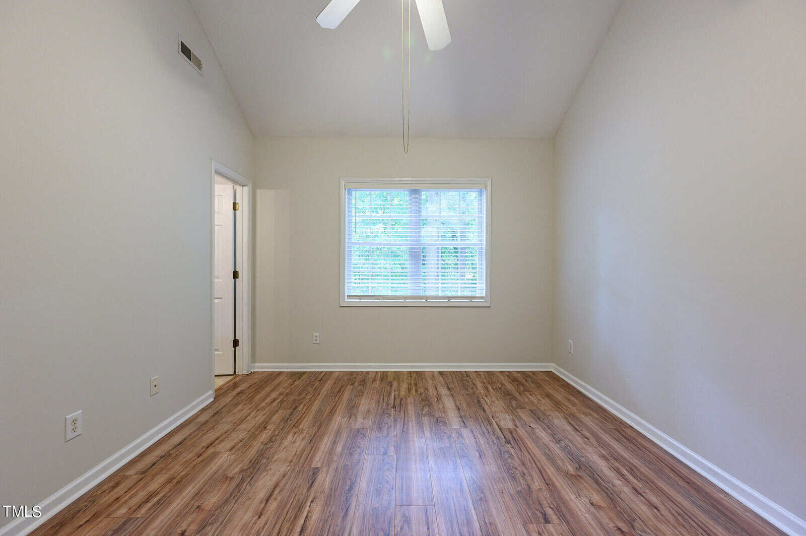 An empty room with light-colored walls, a vaulted ceiling, and a ceiling fan. The hardwood floor leads to a window with blinds, letting in natural light. A doorway to the left reveals a glimpse of another room.