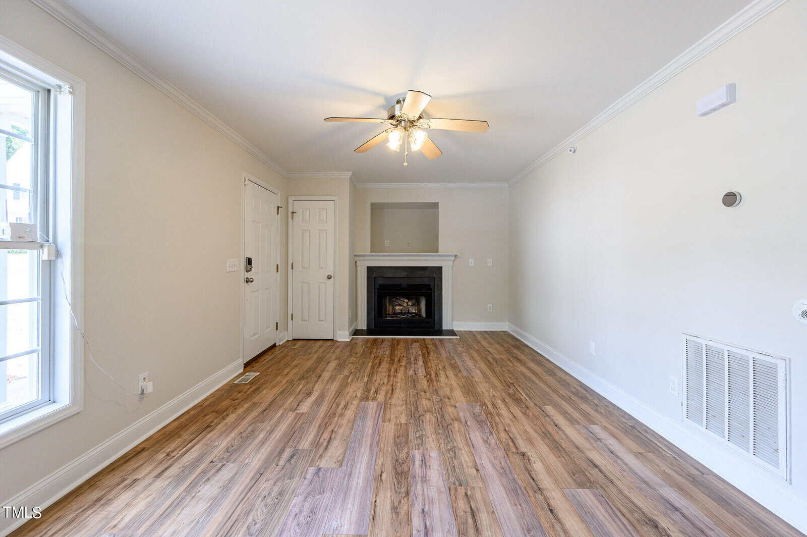 A spacious living room with light wooden flooring, white walls, and a ceiling fan with lights. A fireplace is centered on one wall, and there is a large window on the left, allowing natural light into the room. Multiple doors are visible.